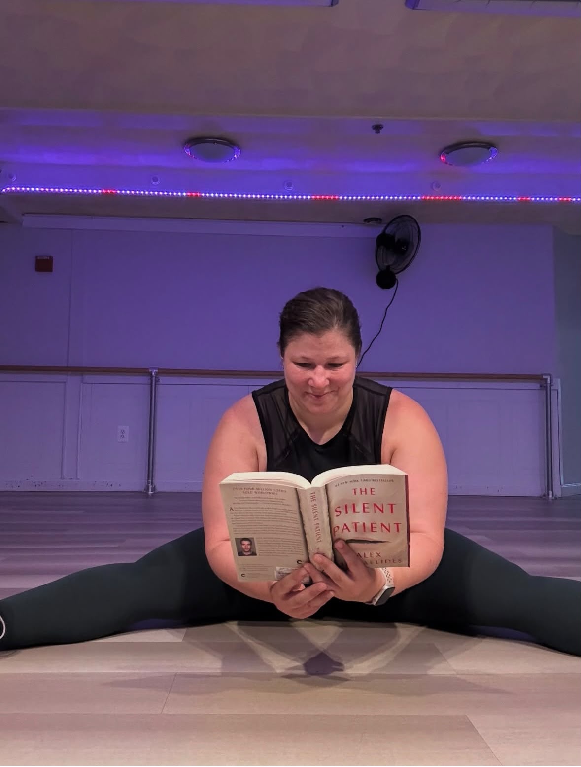 Diana in a seated straddle stretch reading in the purple-lit studio